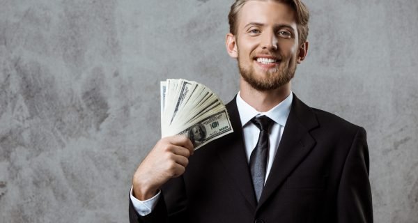 Young successful businessman in suit smiling, holding money over grey background. Copy space.